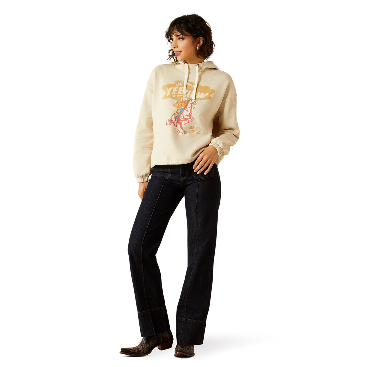 Woman wearing a beige hoodie with graphic design and dark jeans on a white background