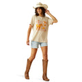 Woman wearing a beige t-shirt with a graphic design, light blue shorts, and brown cowboy boots on a white background.