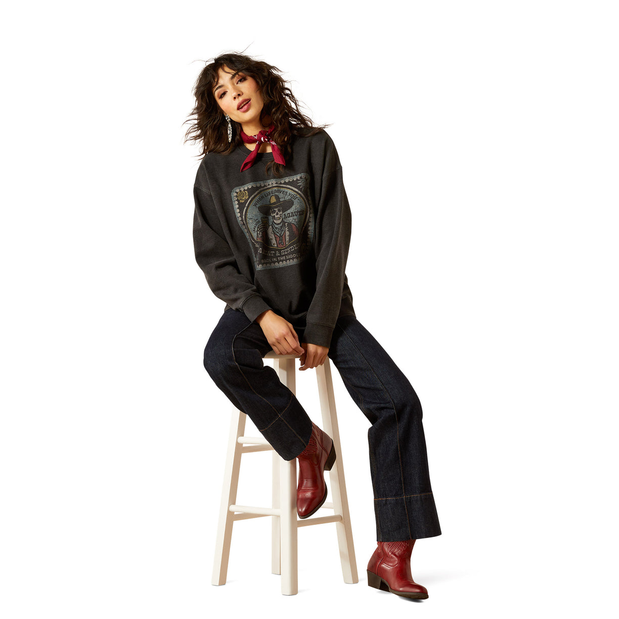 Woman sitting on a stool wearing a dark hoodie with graphic design, jeans, and red shoes on a white background