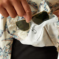 Person holding sunglasses with a floral shirt and white pants in the background