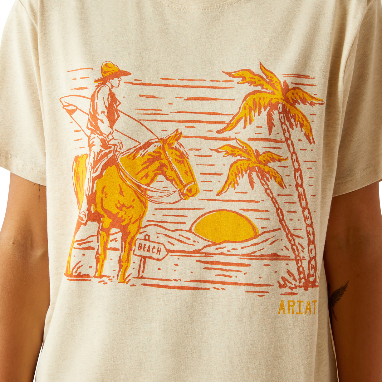 Beige t-shirt with a graphic of a cowboy on a horse, palm trees, and the word 'Ariat' on a beige background.